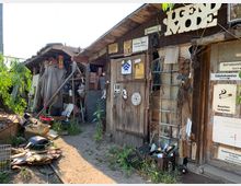 Holzverschlag mit alten, verwitterten Türen und einer Anhäufung von Gegenständen wie Ziegeln, Werkzeugen und Schildern. Die Umgebung ist ungepflegt, mit Gras und verstreutem Müll auf einem sandigen Untergrund.