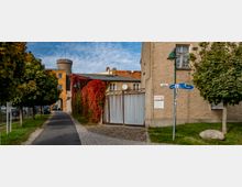 Pflasterweg mit Bäumen und Laternen führt auf ein Tor aus Wellblech zu, das von einer Mauer mit roten, herbstlich gefärbten Kletterpflanzen umgeben ist. Im Hintergrund ein Gebäude aus gelbem Putz mit Turm und Satteldach, rechts ein cremefarbenes Haus mit Straßenschild „Südhaus“ und Wegweisern.