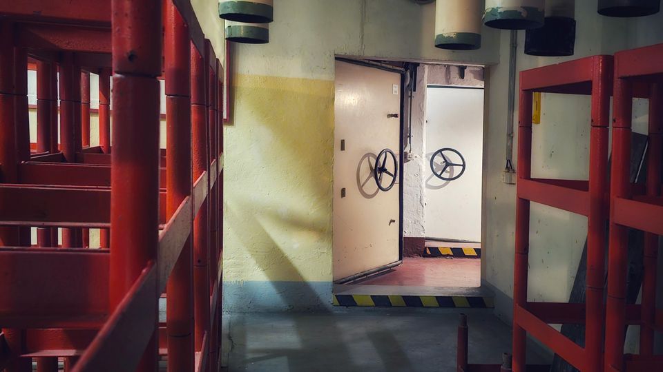 Industriegang in einer technischen Anlage mit roten Metallgeländern und grauem Boden, darüber mehrere große, zylindrische Lüftungs- oder Filterrohre. Am Ende des Ganges stehen schwere weiße Stahltüren mit Handrädern und gelb-schwarzen Warnmarkierungen an den Schwellen.