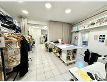 Ein heller Schneiderarbeitsraum mit Fliesenboden, einem großen Zuschneidetisch in der Mitte und Regalen voller Stoffe und Materialien. Links stehen Kleiderstangen mit verschiedenen Kleidungsstücken, während rechts eine Leiter, Schneiderpuppen und Regale mit Ordnern und Büchern zu sehen sind.