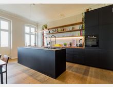 Moderne Wohnküche mit schwarzer Kücheninsel, Arbeitsplatte aus Holz und Edelstahlspüle. Auf der Rückwand befinden sich offene Holzregale mit Büchern, Gläsern und Küchenutensilien, sowie ein Einbaubackofen; der Raum verfügt über hohe Decken, verzierte Stuckelemente und große Fenster mit viel Tageslicht.