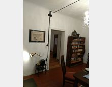 Ein einfacher Innenraum mit weißen Wänden, einem roten Boden und einer Türöffnung, die in einen Raum mit Holzboden führt. An der Wand hängt ein gerahmtes Bild, daneben steht eine kleine Lampe auf einem Regal. Rechts ist ein Holzregal mit Geschirr und Büchern sichtbar, daneben ein Esstisch mit Stuhl.