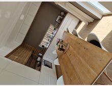 Moderne Küchen- und Esszimmereinrichtung mit einem großen Holztisch und weißen Stühlen. Die Küche verfügt über weiße Einbauschränke, eine Arbeitsfläche in Holzoptik, große Fenster mit Vorhängen und eine Vase mit Blumen auf dem Tisch.