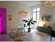 Helles Wohnzimmer mit minimalistischer Einrichtung, bestehend aus zwei Klappstühlen, einem kleinen Tisch mit Kerzen, einer großen Zimmerpflanze und einer grünen Kommode. Im Hintergrund sind eine weiß gerahmte Glastür, abstrakte Wandkunst und rosa Licht, das aus einem angrenzenden Raum scheint, zu sehen.