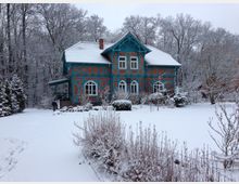 Fachwerkhaus mit roten Ziegeln und türkisfarbenen Holzbalken in einer schneebedeckten Gartenlandschaft, umgeben von winterlichen Bäumen. Ein Mensch steht nahe dem Haus auf dem verschneiten Grundstück.