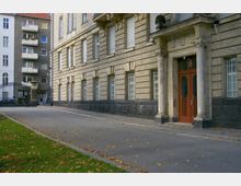 Ansicht einer historischen Fassade eines beige gefärbten Gebäudes mit großen Fenstern, teils vergittert, und einem Eingangsportal mit dekorativen Säulen. Im Vordergrund eine asphaltierte Einfahrt mit herabgefallenen Blättern und angrenzendem Rasen; im Hintergrund stehen mehrstöckige Wohnhäuser.