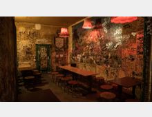 Schmaler Raum mit Tischen und Hockern, dessen Wände vollständig mit Graffiti und Kritzeleien bedeckt sind; schwache Beleuchtung durch rote, hängende Lampen gibt dem Raum eine warme Atmosphäre. Eine grüne Tür am hinteren Ende ist ebenfalls mit Graffiti verziert.