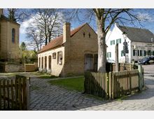 Kleiner historischer Backsteinbau mit Satteldach und Holztüren, umgeben von einem Holzzaun. Links daneben eine Kirche aus hellem Stein und im Hintergrund weitere Gebäude, darunter ein weißes Haus mit grünen Fensterläden.