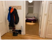 Ein Eingangsbereich mit Holzboden, an dessen Wand eine Garderobe mit mehreren Jacken hängt. Daneben ein Schuhregal mit verschiedener Fußbekleidung. Durch eine Tür ist ein Schlafzimmer mit einem Bett und einem Fenster sichtbar.