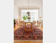 Esszimmer mit einem Holztisch und vier Stühlen auf einem bunten Orientteppich, in einem hellen Raum mit Stuckdecke und großen Fenstern. Im Hintergrund stehen Zimmerpflanzen und Dekorationsgegenstände, die Licht dringt durch die Fenster ein.