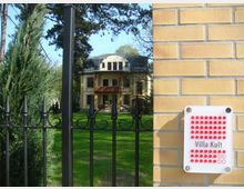 Eine gelbe Villa im Hintergrund mit mehreren Fenstern und einem Mansardendach, umgeben von einem grünen Garten. Im Vordergrund ein schmiedeeiserner Zaun und eine Ziegelmauer mit einem Schild, auf dem "Villa Kult" steht.