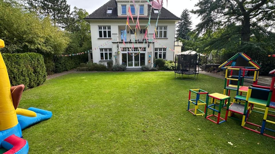 Zweistöckiges Gebäude mit hellen Wänden und dunklem Dach, umgeben von einem Garten mit Rasenfläche. Im Vordergrund stehen Spielgeräte aus bunten Kunststoffstangen, eine Hüpfburg und ein Trampolin, dekoriert mit bunten Wimpelketten.
