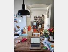 A cluttered home workspace with a wooden table holding a laptop, flowers, books, and various items. In the background, double white doors open into a playroom or study with bookshelves, toys, and colorful decorations, including hanging bunting.