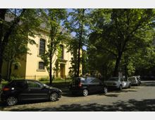 Gelbes Gebäude mit hohen Fenstern im klassischen Stil, umgeben von Bäumen und hinter einem Metallzaun gelegen. Im Vordergrund stehen mehrere parkende Autos entlang einer von Bäumen gesäumten Straße.