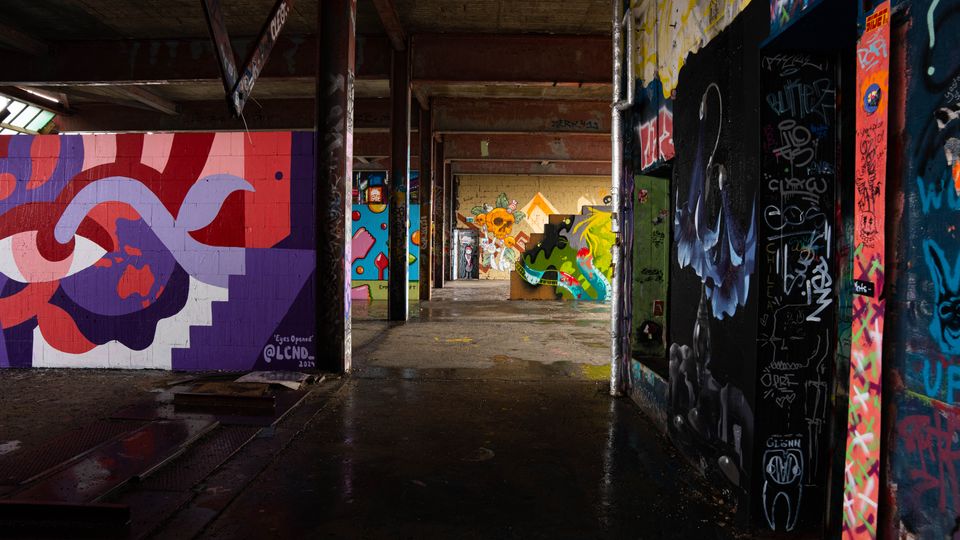 Innenraum einer verlassenen Industriehalle mit Betonboden, Stahlträgern und niedriger Decke, durchzogen von einem Gang. Wände und Pfeiler sind dicht mit großformatigen Graffiti und Street-Art-Motiven in kräftigen Farben bedeckt.