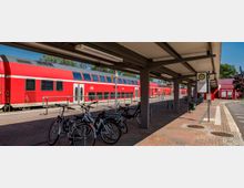 Ein moderner Bahnhof mit einem überdachten Wartebereich, einem roten doppelstöckigen Regionalzug im Hintergrund und Fahrradständern mit mehreren abgestellten Fahrrädern im Vordergrund. Rechts ist ein Schild mit der Aufschrift „Schwedt/Oder Bahnhof“ zu sehen.