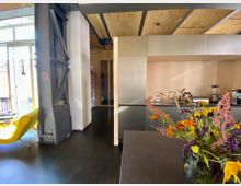 Wohnraum mit industriellem Design, darunter eine freistehende Metallstahlträgerstruktur, eine moderne Einbauküche mit schlichten Fronten und einer Arbeitsfläche. Im Vordergrund steht ein Blumenstrauß mit bunten Blumen auf einem dunklen Tisch, während im Hintergrund ein gelber Sessel und eine Fensterfront zu sehen sind.