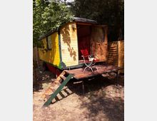 Gelber, hölzerner Bauwagen mit Fenstern und einer kleinen Holzveranda, auf der ein roter Stuhl und ein Tisch stehen. Der Wagen steht in einer lichten, bewaldeten Umgebung und ist über eine schmale Treppe erreichbar.