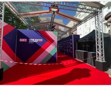 Eine überdachte Veranstaltungsfläche mit rotem Teppich, flankiert von Sponsorentafeln und einem schwarzen Zelt; im Hintergrund sind Gebäudefassaden und Bäume zu sehen. Die Bühne und der Teppich sind von einem transparenten Dach aus Metallträgern eingerahmt.