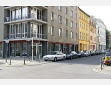 Straßenecke mit mehrstöckigen Wohn- und Geschäftsgebäuden, teils in hellen und farbigen Fassaden gestaltet. Vor den Gebäuden parken Autos entlang des Gehwegs, Fahrräder stehen an Metallpfosten, und Straßenschilder markieren die Kreuzung.