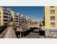 Mehrstöckige Wohngebäude in moderner Architektur mit grauen Fassaden, gelben Akzenten und zahlreichen Fenstern. Im Vordergrund verläuft eine Brücke über eine Straße mit Gehweg und parkenden Autos; an einer Hausfassade ist ein Wandgemälde sichtbar.