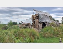 Eine einfache Holzhütte mit unregelmäßigem Dach aus Planen steht inmitten einer natürlichen, von Gras und Sträuchern bewachsenen Landschaft. Die Konstruktion besteht aus unverarbeiteten Holzästen und Brettern, umgeben von freiem Himmel und vereinzelten Bäumen im Hintergrund.