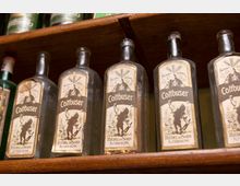 Nahaufnahme eines Holzregals mit mehreren Flaschen des Getränks „Cottbuser“, die ein markantes Etikett mit einer Illustration und der Aufschrift „Riedel & Sohn, Altdöbern“ tragen. Im Hintergrund stehen weitere grüne Glasflaschen.