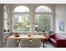 Essbereich mit einem Holztisch, umgeben von weißen und roten Stühlen, vor drei großen Rundbogenfenstern, die einen Blick auf einen Garten und ein Backsteingebäude bieten. Im Raum stehen Pflanzen, eine rote Sofa-Ecke und eine Hängelampe über dem Tisch.