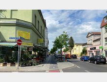 Straßenszene mit Blumengeschäft auf der linken Seite eines grün gestrichenen Gebäudes, dessen Fassade mit Pflanzen dekoriert ist. Rechts der Straße stehen geparkte Autos, zwischen Bäumen und weiteren Gebäuden in hellen Farben; ein Fahrradweg führt parallel zur Fahrbahn.