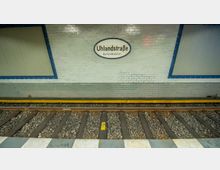 U-Bahn-Station mit weiß gefliester Wand, einem ovalen Schild mit der Aufschrift „Uhlandstraße Kurfürstendamm“ und blau umrandeten Wandpanelen. Im Vordergrund sind Schienen und ein gelber Sicherheitsstreifen sichtbar.