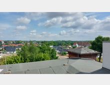 Blick über eine Wohnsiedlung mit einzelnen Häusern und roten Dächern, umgeben von Bäumen und Grünflächen. Im Vordergrund sind Dächer eines Gebäudes sichtbar, im Hintergrund erstreckt sich die Landschaft unter einem leicht bewölkten Himmel.