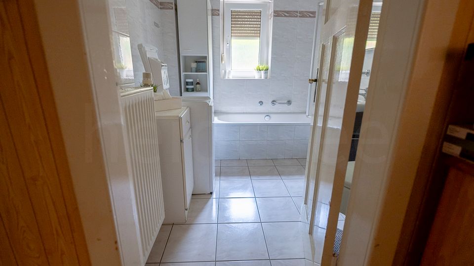 Blick durch eine Tür in ein hell gefliestes Badezimmer mit großer Badewanne an der Stirnwand und einem Fenster darüber. Links stehen weiße Badmöbel und ein hoher Schrank, rechts ist eine weitere Tür zu einem angrenzenden Bereich sichtbar.