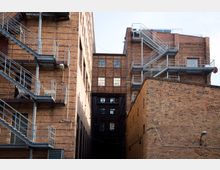 Backsteingebäude mit industriellem Stil, versehen mit Außentreppen aus Metall. Der schmale Innenhof wird von hohen Fassaden flankiert, die teils durch Fenster und Verbindungselemente oberhalb des Durchgangs geprägt sind.