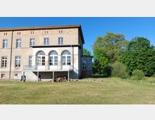 Historisches Gebäude mit zwei Stockwerken, großflächigen Fenstern und heller Fassade, ergänzt durch einen überdachten Balkon mit Rundbogenfenstern. Im Vordergrund erstreckt sich eine gepflegte Wiese, im Hintergrund sind große Bäume sichtbar.