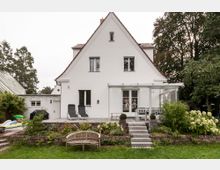 Ein weißes Einfamilienhaus mit Satteldach und großen Fenstern, betrachtet von der Gartenseite. Im Vordergrund ein gepflegter Garten mit Steinweg, Terrasse mit Sitzmöbeln und Liegestühlen, sowie umliegenden Bäumen und Sträuchern.