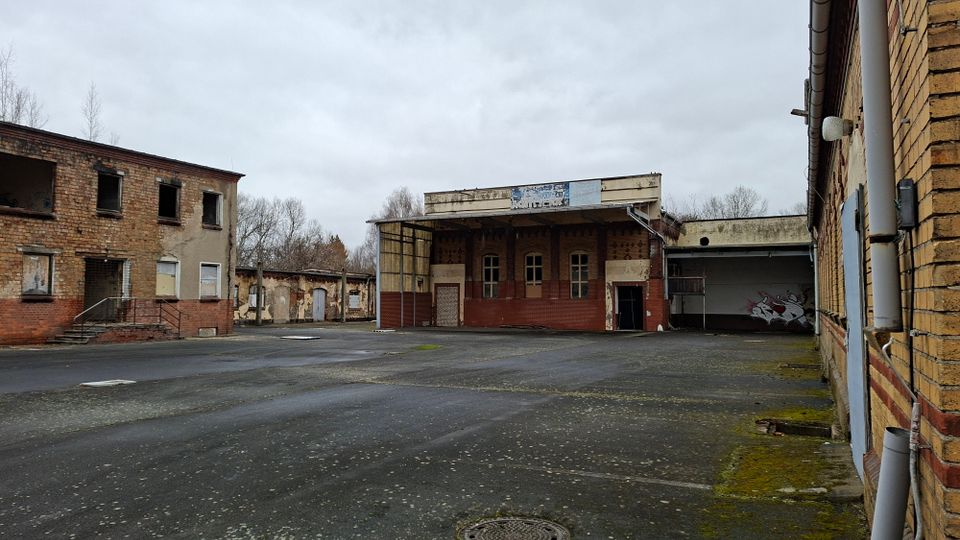 Verlassener Industriehof mit weitläufiger Asphaltfläche, umgeben von mehrstöckigen Backsteingebäuden mit beschädigten Fenstern und abgeblätterter Fassade. In der Mitte steht ein flacher Bau mit überdachtem Eingangsbereich und großen Fenstern, daneben eine offene Halle mit Graffiti.