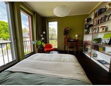 Helles Schlafzimmer mit olivgrünen Wänden, großem Bett im Vordergrund und bodentiefen Fenstern mit Balkonblick ins Grüne. Rechts stehen ein langes Bücherregal und ein kleiner Schreibtisch; im Hintergrund ein roter Sessel, Pflanzen und eine runde Deckenlampe.