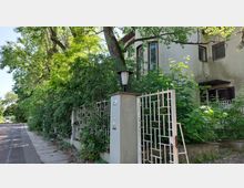 Wohnhaus mit beiger Fassade, umgeben von dichter Vegetation und einem weißen Metallzaun. Im Vordergrund steht ein Pfeiler mit einer Laterne und einer Hausnummer, daneben ein Gehweg unter Bäumen.