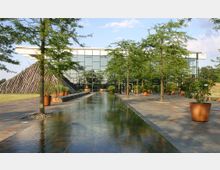 Modernes Gebäude mit einer Fassade aus großen Glasfenstern, davor eine gepflasterte Fläche mit einer schmalen Wasserfläche in der Mitte und mehreren in Töpfen gepflanzten Bäumen. Links ist ein pyramidenförmiges Gebilde aus gestapelten Holzbalken zu sehen.