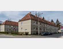 Mehrfamilienhaus in einem Wohngebiet mit hellbeigen Fassaden und roten Ziegeldächern. Die Gebäude stehen an einer Straßenecke mit parkenden Autos, Straßenlaternen und begrenzenden Hecken.