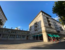 Großer Platz vor einem mehrstöckigen Gebäude mit heller Fassade und rechteckigen Fenstern. Im unteren Bereich des Gebäudes befinden sich grüne Markisen über Schaufenstern und teils aufgestellten Stühlen und Tischen, während Statuen auf dem Dach des angrenzenden Gebäudeteils zu sehen sind.