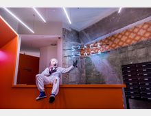 Ein Innenraum mit einer Mischung aus moderner und industrieller Gestaltung, sichtbaren Betonwänden und einer Wanddekorleiste mit orangefarbenem Retro-Muster. Ein weiß gekleideter Raumanzug-Dummy sitzt auf einer orangefarbenen Theke, während darüber ein Neon-Schriftzug mit den Worten „Space Peace“ leuchtet.