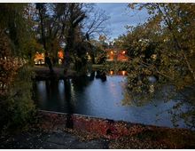 Blick über einen kleinen Teich in einem Park bei Dämmerung, umgeben von hohen Bäumen mit herbstlichem Laub. Im Hintergrund spiegeln sich beleuchtete Gebäude und Straßenlampen im Wasser, vorne ist eine niedrige, überwachsene Ufermauer zu sehen.