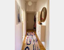 Schmaler, heller Flur mit Holzboden, einem bunten, gemusterten Teppich und einer langen Garderobe mit weißen Türen an der rechten Wand. An der Wand hängt ein runder Spiegel, daneben stehen eine Vase mit Blumen und eine Zimmerpflanze. Links befindet sich ein gerahmtes Bild, die Deckenbeleuchtung ist kugelförmig.