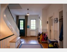 Heller Hausflur mit Holzfußboden, Treppe mit weißem Geländer und grünem Handlauf sowie einer grünen Eingangstür. Neben einem Fenster mit Vorhängen stehen eine kleine Bank mit Garderobe, ein Tisch und eine Deckenleuchte; rechts sind Innentüren und Jacken an Haken.