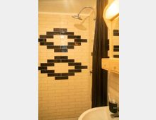 Duschbereich eines Badezimmers mit beige und schwarzen Kacheln im rechteckigen Design, einer Regendusche und einem schwarzen Duschvorhang. Im Vordergrund ein Waschbecken mit Zahnbürstenbehälter auf der Ablage.