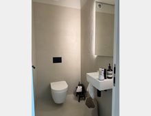 Modernes Badezimmer mit heller, minimalistischer Gestaltung, beige Fliesen an Wänden und Boden. Ein wandmontiertes WC, ein kleines Waschbecken mit Accessoires sowie ein Hocker mit Toilettenpapier und ein Korb sind sichtbar.