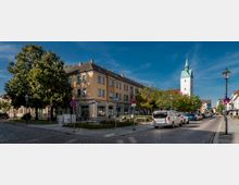 Straßenansicht mit einem gelben Wohn- und Geschäftsgebäude auf der linken Seite, einer Grünfläche mit Bäumen und Sitzbänken sowie einer Kirche mit weißem Turm und grünem Spitzdach im Hintergrund. Autos parken entlang der gepflasterten Straße auf beiden Seiten, und Laternen sowie Verkehrsschilder sind sichtbar.