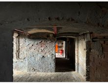 Dunkler, verwitterter Kellerraum mit freiliegenden Backsteinwänden und gewölbter Decke. Im Vordergrund sind rostige Rohre und abgenutzte Steinböden zu sehen, während im Hintergrund ein orangefarbener Schrank und ein roter Stuhl stehen.