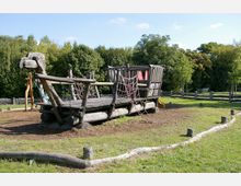 Ein hölzernes Klettergerüst in Form eines Schiffs mit Netzseilen und Rutschen, umgeben von Rasen und Bäumen in einem Spielplatzbereich. Im Hintergrund stehen Holzzäune und dichter Baumbewuchs.